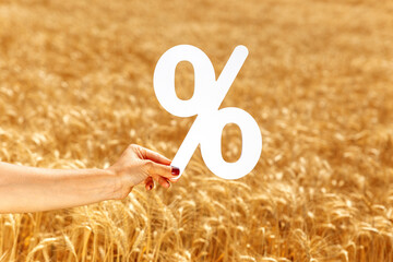 Female hand holding a large white percent symbol over a golden wheat field, representing...