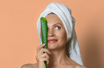 Woman wrapped in white towel covering one eye with aloe leaf and smiling. Peach studio background.
