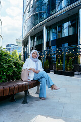 Muslim woman relaxing on bench with handbag near office