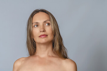 Dreamy blonde with bare shoulders looks aside against gray. Cosmetic procedures, rest and body care.