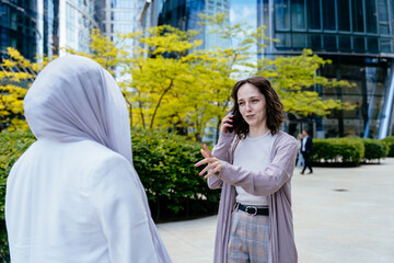 Woman talking on phone in front of Muslim friend outdoors