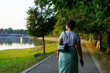 Woman walking along a lakeside path surrounded by trees during late afternoon in a serene setting