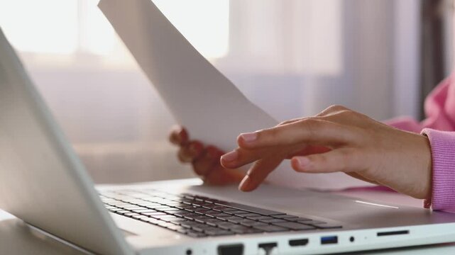 A close-up shot of hands typing on a laptop keyboard while holding a document or paper, with soft background light.