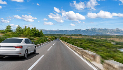 A white car speeds down an open highway through a scenic mountain landscape under a bright blue sky...
