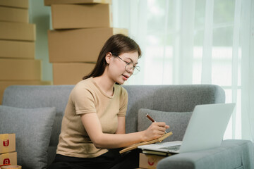 A young Asian woman sits on a sofa, writing a message on a package for delivery, surrounded by...