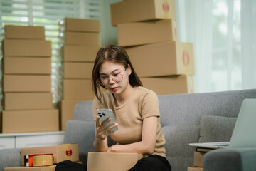 Stressed Asian woman sits on a sofa, surrounded by packages. She holds her head in frustration as...