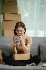 Stressed Asian woman sits on a sofa, surrounded by packages. She holds her head in frustration as...