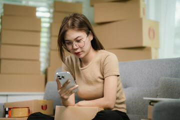 Stressed Asian woman sits on a sofa, surrounded by packages. She holds her head in frustration as...