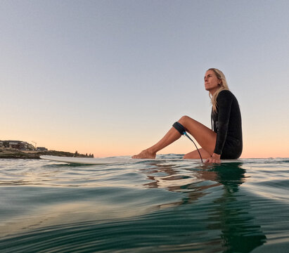 Woman sitting on surfboard floating on glassy water with leg up on board and sunrise sky