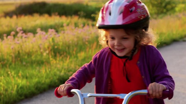 A young girl with a pink helmet smiling at the camera as she rides her bicycle on a paved path next to a field of green grass and purple wildflowers at sunset.