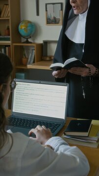 Vertical of student girl working on laptop sitting at desk with unrecognizable nun talking in front of class holding Bible and rosary