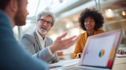 Three people are sitting at a table with a laptop open in front of them. They are discussing something important, and the laptop screen shows a graph. Scene is serious and focused