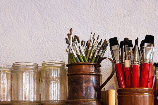 Closeup of assorted paintbrushes stored in a vintage copper pitcher and empty glass jars against a textured wall
