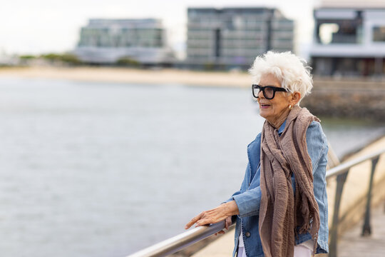 elderly woman standing at the waterfront leaning on railing with apartment building in background