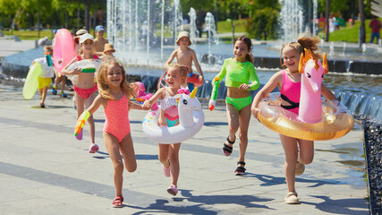 Kids joyfully sprint with pool inflatables by park fountains. Concept of toy brand marketing image, swimwear catalog, seasonal children product promotion