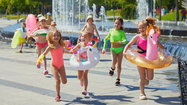 Kids joyfully sprint with pool inflatables by park fountains. Concept of toy brand marketing image, swimwear catalog, seasonal children product promotion