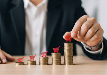 A professional businessman in a black suit is arranging small red shopping cart icons on top of stacks of gold coins, concept growth in sales or investments.