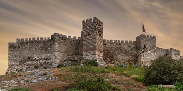Ayasuluk Castle, Selcuk, Turkey. Ancient stone fortress walls, battlements, and a tower with the Turkish flag, seen from a rocky, vegetated hill against a cloudy sky - Powered by Adobe