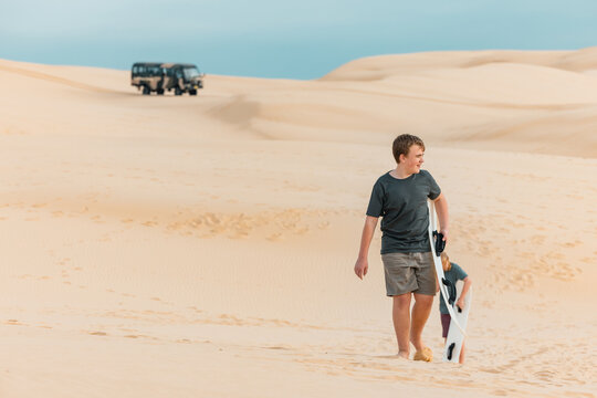 Kids carrying sand boards across dunes with off road tour bus in background at Birubi Beach