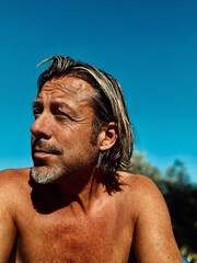 Tanned man with half long blond hair and grey stubble beard, shirtless outdoor in summer sun under clear blue sky.