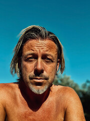 Tanned man with half long blond hair and grey stubble beard, shirtless outdoor in summer sun under clear blue sky.