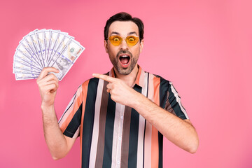 Excited man holding a fan of cash against a pink background, expressing joy and success in casual...