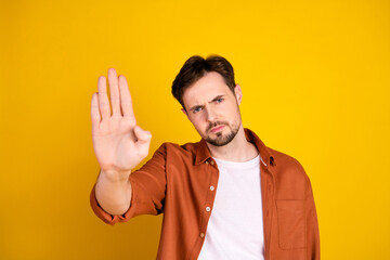 Young man in orange shirt showing stop gesture against vibrant yellow background portraying...