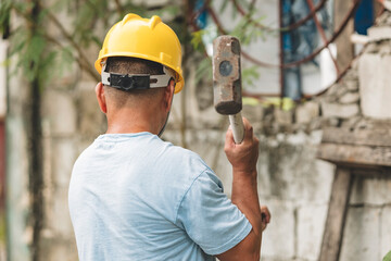 Rear view of Southeast Asian man in hard hat swinging sledgehammer against crumbling wall during a...