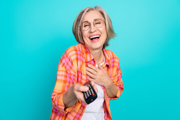 Joyful senior woman in stylish checkered shirt holding remote control against teal background,...