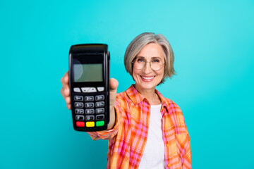 Stylish elderly woman showing a card payment terminal against a teal background for advertising or...