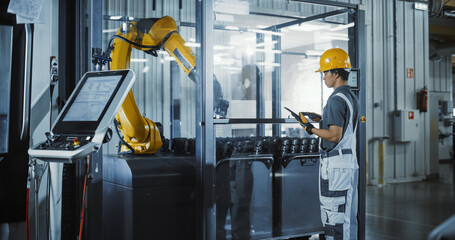 Asian Male Engineer Using Laptop Computer to Control a Robotic Arm at a Metal Components Production Factory. Young Man Working on Manufacturing of Innovative Parts for Artificial Intelligence Systems