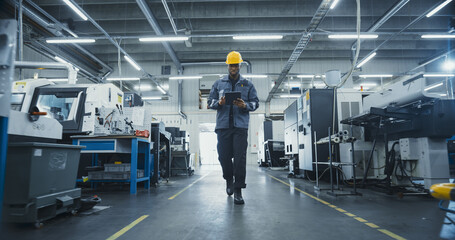 African American Male Worker in a Yellow Hard Hat Walks Through a Modern Factory, Inspecting Equipment and Recording Data on a Tablet Computer. Facility is Equipped with Advanced Machinery