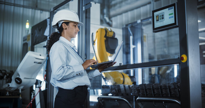 Industrial Engineer at Work. Hispanic Female Using Laptop Computer to Monitor a Robotic Arm in a High Tech Metal Factory, Ensuring Precise Automation and Efficiency in Production Process