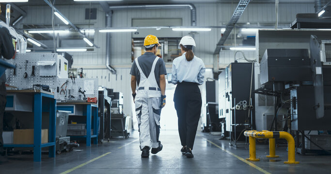 Diverse Female and Male Technicians Walking Through a Modern Factory, Discussing Work. Young Man Wearing a Yellow Hard Hat and Overalls, Manager Dressed in Business Attire. Footage From the Back