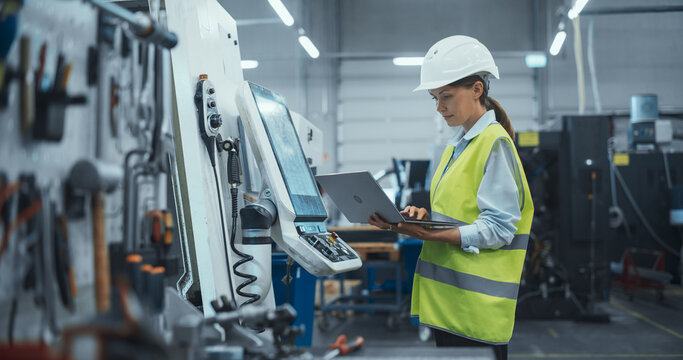 Female Machinery Technician Using Laptop Computer and Programming a CNC Machine at an Advanced Metal Production Facility. Young Woman in Work Gear Setting Up Manufacturing Process in a Factory