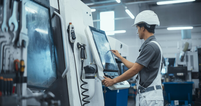 Asian Male Engineer in a White Hard Hat Operating a CNC Milling Machine in a Modern Factory. Young Technician Inserting Parameters on a Touch Screen Display, Automating Production Line