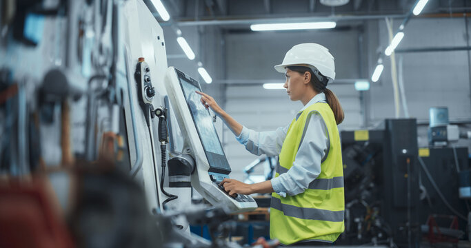 Female Machinery Technician Using Laptop Computer and Programming a CNC Machine at an Advanced Metal Production Facility. Young Woman in Work Gear Setting Up Manufacturing Process in a Factory