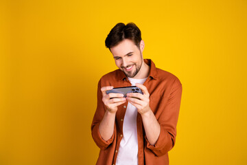 Fototapeta premium Young man enjoying leisure gaming on smartphone while smiling against a vibrant yellow background in stylish attire