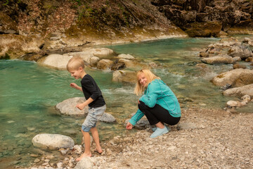 Young-looking grandmother with grandson on the shore of a mountain river. Throwing stones into emerald clear water. Peaceful nature, bonding moments, summer day outdoors. 