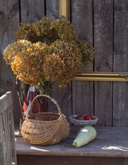 In the garden shed, a composition with old, vintage things - a chair, a table and a basket on it, zucchini, red peppers in a bowl, dried flowers in a metal can.