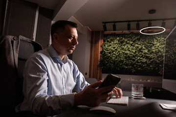Businessman in a light shirt types on keyboard and phone in a dark elegant office. Evening light,...