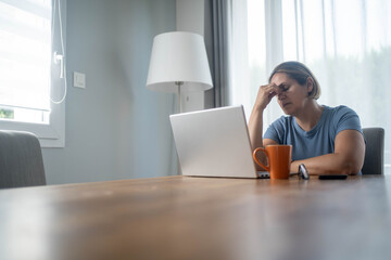 Stressed woman managing bills and rising living costs at home, sitting at desk in front of laptop,...
