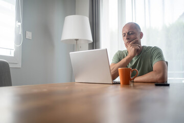 Mature man thinking while looking at laptop screen at home, reflecting on ideas, focused and calm...