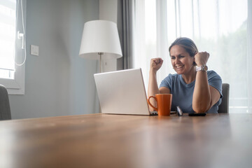 Excited woman raising fists in joy, celebrating success with laptop at home, concept of happiness,...