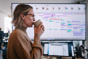 A middle-aged Caucasian woman juggles deadlines while sipping coffee at her standing desk, managing emails, video meetings, and detailed Gantt charts for a high-stakes corporate rollout