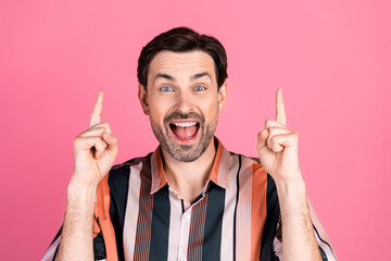 Cheerful young man pointing upward with both hands against a vibrant pink background showcasing...