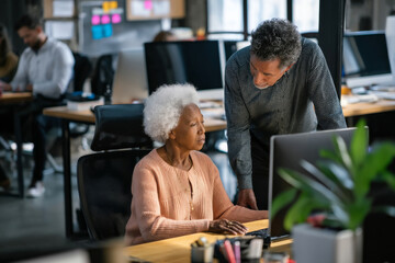An elderly Black woman checks ergonomic seating arrangements in the workspace, consulting with a wellness coach to ensure every employee has optimal support for their health and productivity