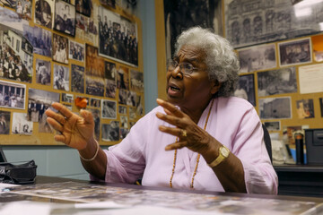 An elderly Black woman hosts a history walk through the office, telling stories from the company's past as photos and memorabilia line the hallways, bridging generations and legacy