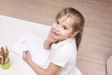 Naklejka premium Caucasian female child smiling and drawing at table with colored pencils in bright room.