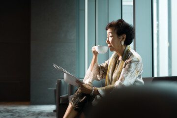 An elderly Asian woman enjoys a moment in a bright office lounge, sipping coffee while reading, wearing a silk blouse and exuding wisdom and calm through the soft window light.
